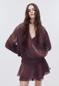 Bruine doorschijnende blouse met oversized lange mouwen en brede tailleband, gecombineerd met een korte, gelaagde rok. Gladde textuur, V-hals ontwerp.