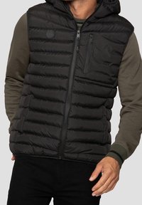 Doudoune sans manches noire avec capuche, présentant un motif matelassé, une fermeture éclair frontale et une poche poitrine. Portée sur une chemise à manches longues verte.