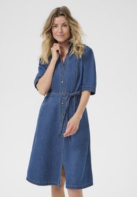 Ženska z blond lasmi, oblečena v modro denim midi obleko z gumbi in zavezanim pasom, stoji ob svetli enobarvni ozadju.