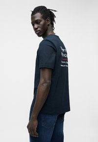 Fiatal férfi sötét fonott hajjal, navy kék Tommy Hilfiger pólót és kék farmert visel, fehér háttér előtt hátrafordulva néz.