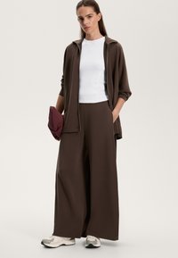 Hnedá prešívaná bunda s tónmi, biely priliehavý top a široké hnedé nohavice. Obsahuje vínovú tašku a svetlé tenisky so šedým vzorom.