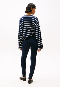 Kék-fehér csíkos, hosszú ujjú felső, slim-fit sötét farmer és fekete lapos cipők. Modell hátulról, sima fehér háttér előtt.