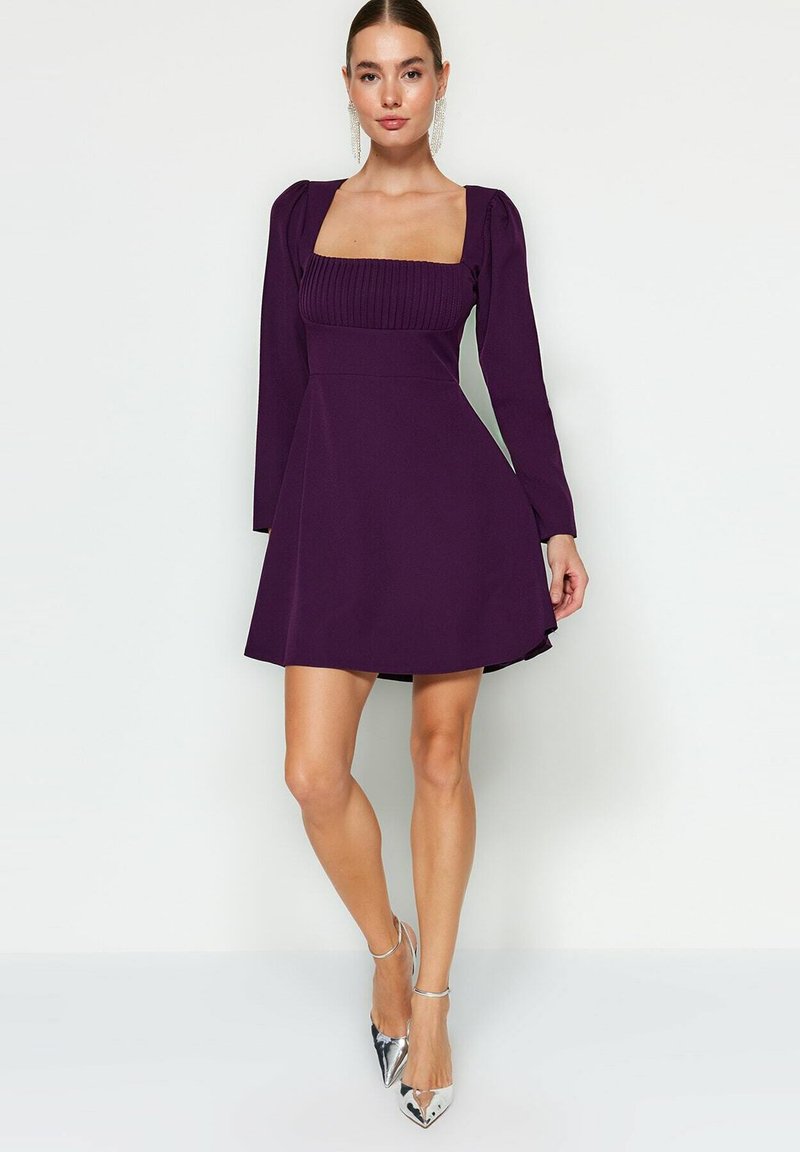 Robe violette avec un décolleté carré, un corsage texturé, des manches bouffantes et une jupe évasée. Associée à des talons aiguilles argentés.