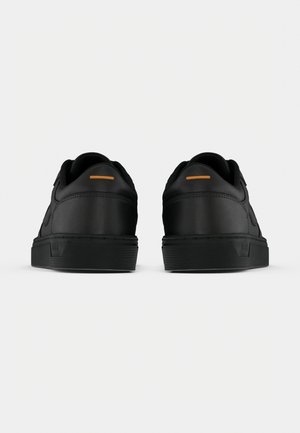 Paire de baskets basses noires vues de dos avec un détail orange minimal sur la languette du talon et des semelles en caoutchouc épaisses.