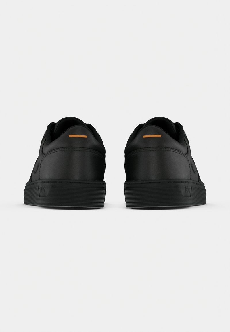 Paire de baskets basses noires vues de dos avec un détail orange minimal sur la languette du talon et des semelles en caoutchouc épaisses.