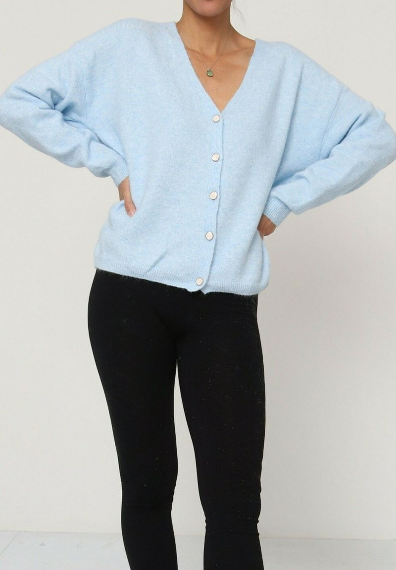 Cardigan bleu clair à boutons, avec un col en V, coupe oversize et poignets côtelés. Texture douce, porté par-dessus des leggings noirs.