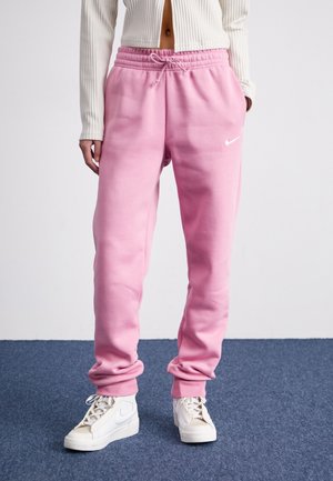 Person trägt hellrosa Nike-Sweathosen mit elastischen Bündchen und weiße Nike High-Top-Sneaker, steht auf blauem Teppich.