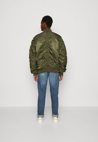 Olive green bomber jacket with ribbed cuffs and collar, featuring a smooth, shiny texture and gathered detailing along the back seam, worn over blue jeans.