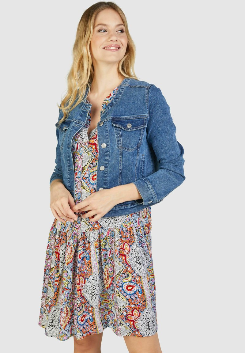 Die Jeansjacke verfügt über ein klassisches Design mit vorderen Taschen und wird mit einem bunten, gemusterten Kleid in Rot-, Blau- und Weißtönen kombiniert.