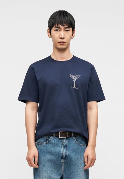 Μπλε σκοτεινό βαμβακερό μπλουζάκι με εμπρόσθιο γραφικό τύπωμα, στρογγυλή λαιμόκοψη και κοντά μανίκια, συνδυασμένο με ανοιχτό μπλε τζιν παντελόνι και μαύρη ζώνη.