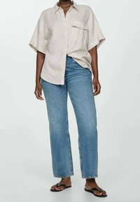 Personne portant une chemise boutonnée à manches courtes de couleur beige clair, un jean bleu à jambe droite et des sandales plates noires, se tenant devant un fond uni.