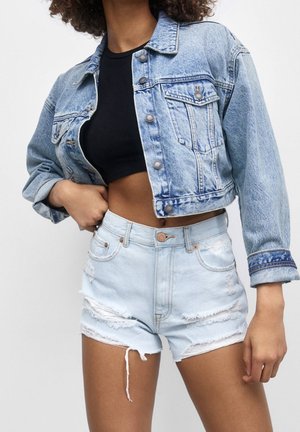 Chaqueta de mezclilla azul claro con un diseño recortado, botones plateados y un leve desgastado. Combinada con pantalones cortos de tiro alto desgastados azul claro.