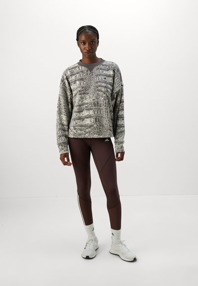 Kvinne som står mot hvit bakgrunn iført grå mønstrete hettegenser, mørkebrune leggings med logo, hvite sokker og hvite joggesko.
