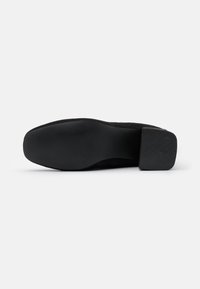 Zapato de ante negro con punta redondeada, tacón bajo y suela texturizada. La parte superior es lisa, consistente en color y diseño.