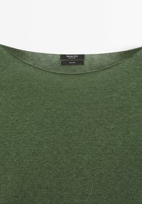 Encolure d'un pull en maille verte avec une étiquette noire portant la mention "Massimo Dutti, Fabriqué en Chine, 100 % lin" sur un fond blanc.