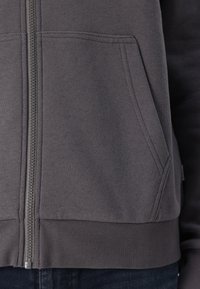 Felpa grigia con zip, caratterizzata da un ampio orlo a coste e polsini, con una texture morbida e tasche frontali. La zip si estende centralmente lungo la parte anteriore.