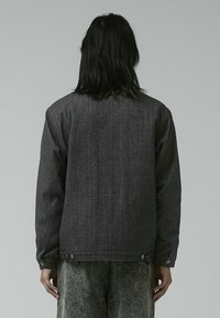 Chaqueta de denim gris oscuro con un corte relajado, que cuenta con botones a presión en el dobladillo y un acabado texturizado. La vista trasera muestra elementos de diseño mínimos.