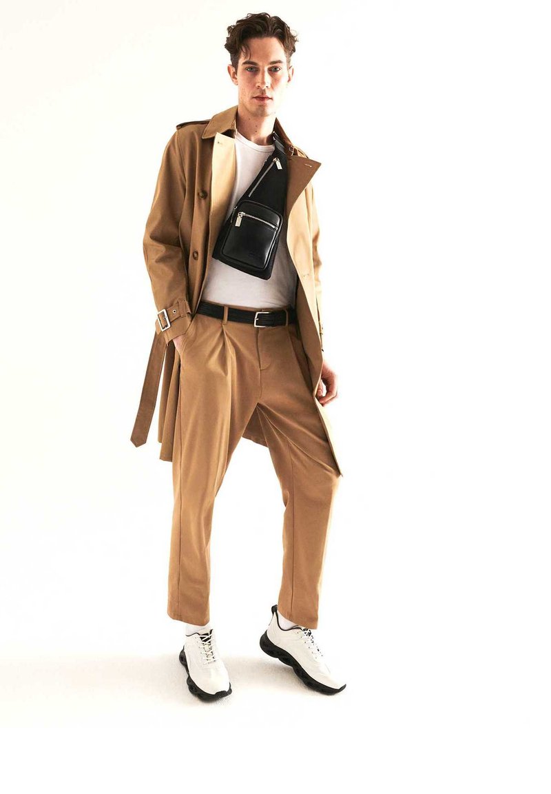 Trench coat tan sopra una maglietta bianca, abbinato a pantaloni beige e sneakers bianche. Presenta anche una piccola borsa a tracolla nera.