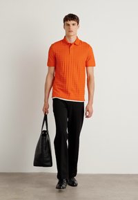 Polo laranja texturizado, calças pretas e bolsa preta. Modelo em pé contra um fundo simples, exibindo um design limpo.