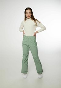 Helle grüne Skihosen mit geradem Schnitt, verstellbarem Bund und zwei frontalen Taschen. Kombiniert mit einem weißen gerippten Rollkragenpullover und Sneakers.