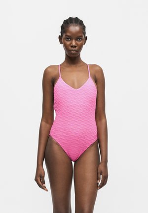 Femme debout face à l’objectif portant un maillot de bain une pièce rose texturé avec de fines bretelles sur un fond blanc uni.