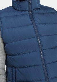 Pälsad marinblå ärmlös väst med hög krage, vertikal quiltad mönster och dragkedja fram. Har grå tygdetaljer på sidan.