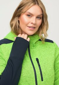 Giacca in fleece verde con accenti blu scuro, colletto alto e tasca sul petto con zip. Tessuto a coste, maniche aderenti con aperture per i pollici.