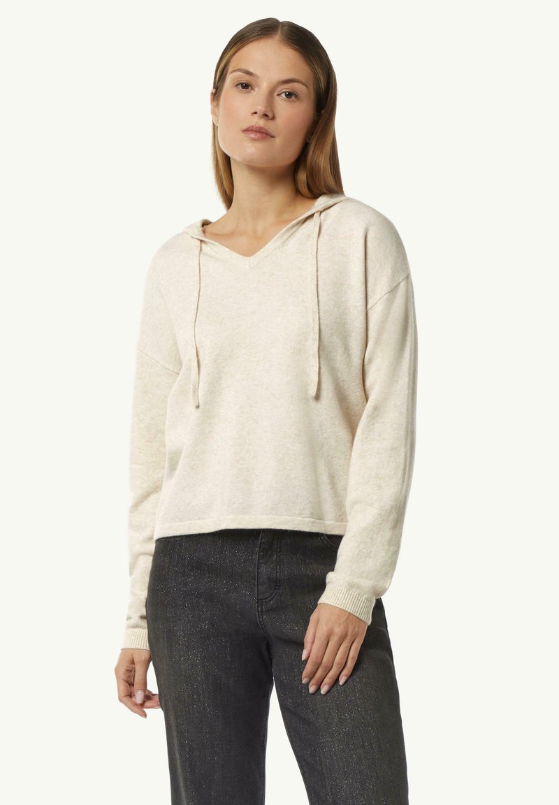 Cremfarbener, kurzer Hoodie mit V-Ausschnitt, Kordelzug und gerippten Bündchen. Weiche Textur mit lockerer Passform, kombiniert mit dunklen Jeans.