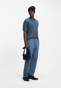 Miehen asu koostuu teksturoidusta tummansinisestä t-paidasta, vaaleansinisistä ohutraitaista housuista, mustista jalkineista ja pienestä mustasta olkalaukusta.