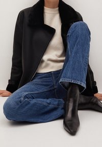 Personne assise portant une veste en peau lainée noire, un pull crème, un jean bleu et des bottes noires à bout pointu sur un fond blanc uni.
