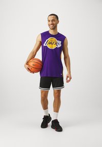 Lila brezrokavni dres s grafiko "Los Angeles Lakers", črne kratke hlače z belimi detajli, drži košarkaško žogo, obut v črne športne copate.