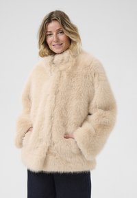 Svetainio aksominio audinio bežis paltas su aukštu apykakle, plačiomis rankovėmis ir šoninėmis kišenėmis. Minkšta tekstūra su puriu apdaila ir besiūle konstrukcija.