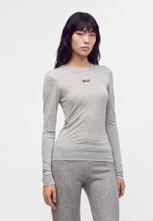 Donna con lunghi capelli scuri che indossa una maglia a maniche lunghe grigio chiaro con un piccolo logo nero "Lacoste" e pantaloni grigi coordinati.