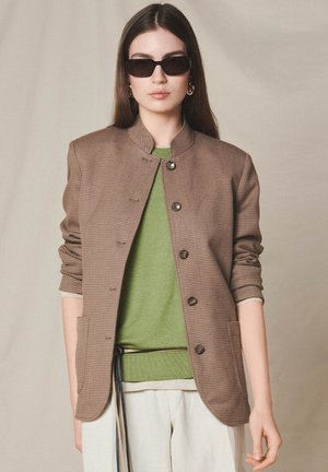 Kvinde iført mørke solbriller, en brun tekstureret knapjakke over en grøn sweater og lyse bukser foran en ensfarvet baggrund.