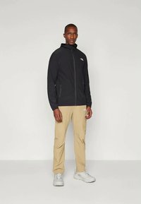 Fekete zipzáras dzseki kapuccsal és logóval, bézs nadrág és fehér sportcipő. Az sima anyag és a testhezálló kivitel fokozza a funkcionalitást.