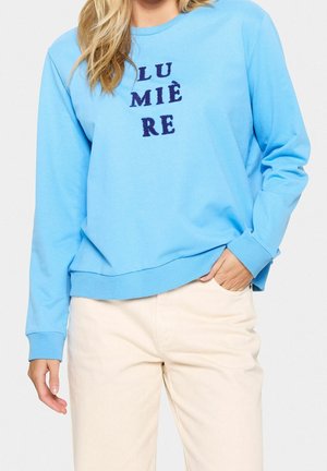 Kvinde iført lyseblå sweatshirt med mørkeblå bogstaver, der staver "LUMIÈRE", og lyse beige bukser, med hånden i lommen.