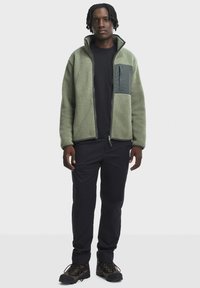 Fleece bunda v tlmenej zelenej farbe s tmavozeleným zipsovým vreckom na hrudi, skombinovaná s čiernymi nohavicami a textúrovanými čiernymi turistickými topánkami.