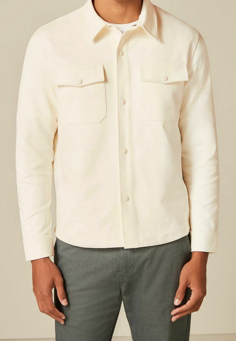 Homme portant une chemise crème à manches longues boutonnée avec deux poches poitrine et un pantalon gris foncé texturé, debout devant un fond uni.