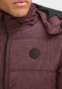 Bordeaux farvet pufferjakke med struktureret finish, sorte accenter på skuldrene, lynlås foran og et rundt logo-patch med teksten "DENIM BLEND."