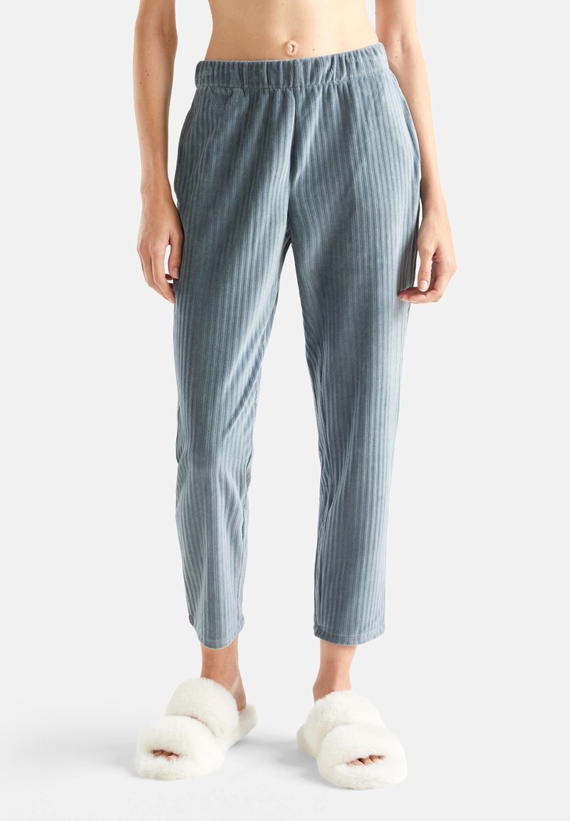 Blå ribbade loungbyxor som bärs av en person som står barfota i vita fluffiga tofflor mot en enfärgad bakgrund.