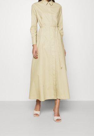 Woman wearing a long beige shirt dress with a waist tie and long sleeves, paired with white open-toe heeled sandals on a plain background.