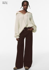 Maglione lavorato a maglia crema con spalle scoperte, abbinato a pantaloni marroni a vita media e gamba larga. Taglio oversize e tessuto texturizzato distintivi.
