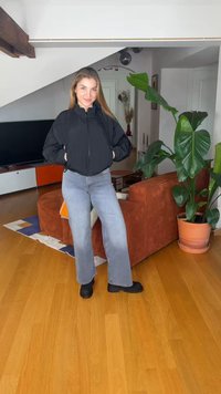 Jauna sieviete melnā žaketē, pelēkos džinsu biksēs un melnos zābakos stāv viesistabā ar koka grīdu un lielu zaļu podā iestādītu augu.