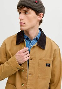 Ung mand iført en brun kordfløjlsjakke med krave over en denimskjorte, justerer jakken, og en brun strikhue med et rødt prymærke.