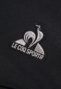 Tessuto nero con un logo ricamato che presenta un gallo stilizzato e il testo "LE COQ SPORTIF" in argento.