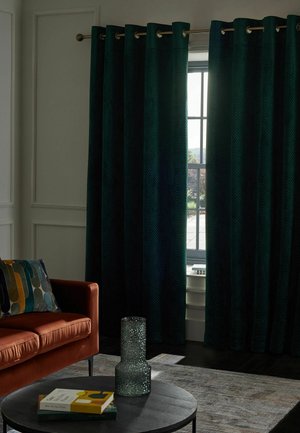 Dunkelgrüne schwere Vorhänge, die ein Fenster teilweise bedecken, neben einem rostfarbenen Sofa mit einem mehrfarbigen Kissen und einem runden Tisch mit Büchern und einer strukturierten Vase.