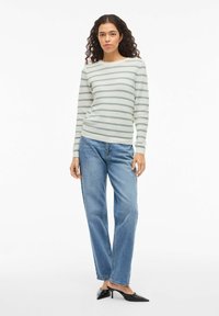 Femme aux longs cheveux bouclés portant un pull rayé blanc et vert, un jean bleu et des talons pointus noirs, debout sur un fond blanc.