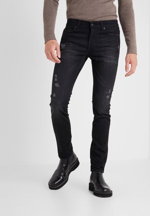 Man draagt zwart slim-fit distressed jeans, een bruin shirt met lange mouwen en zwarte leren enkellaarzen, staand tegen een effen achtergrond.