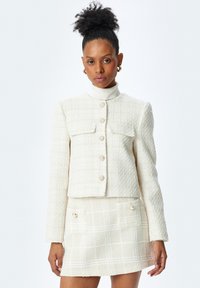 Giacca bianca in tweed con due tasche frontali e finitura testurizzata, dotata di chiusure con bottoni in perla. Abbinata a una gonna mini coordinata.