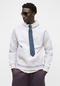 Νεαρός άνδρας που φορά γυαλιά ηλίου, ανοιχτό γκρι φούτερ με κουκούλα και μπλε γραβάτα, καρό πουκάμισο από κάτω και σκούρο μοβ παντελόνι, στέκεται με τα χέρια στις τσέπες.